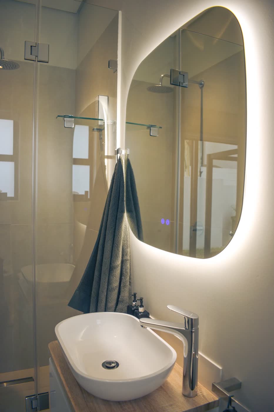Modern bathroom interior with a backlit irregular-shaped mirror, vessel basin on a timber counter and frameless glass shower