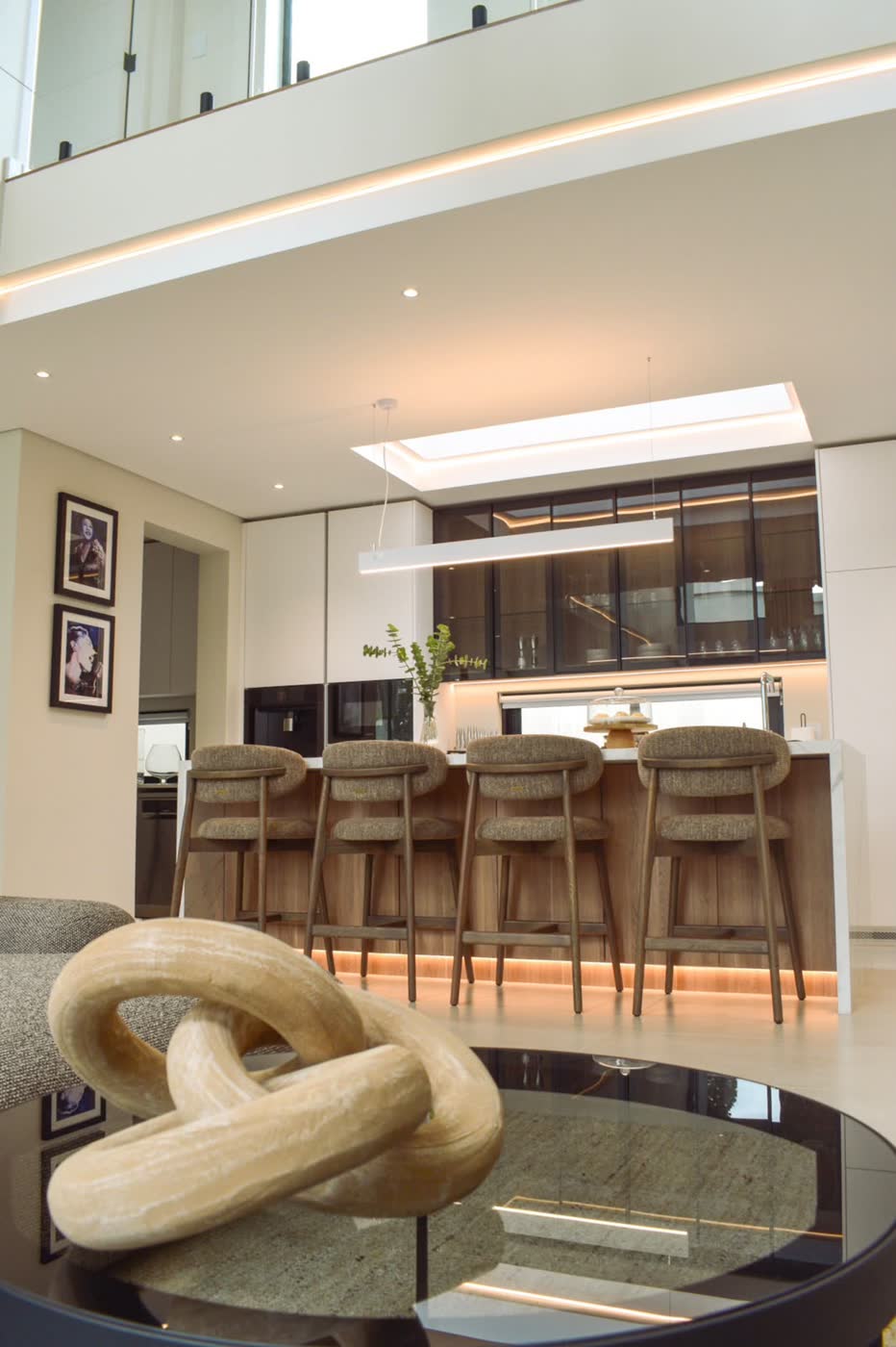 Double-volume open-plan kitchen and living space with a timber breakfast bar, upholstered bar stools and warm cove lighting