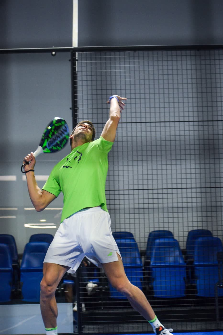 Padel world number one Juan Lebrón Chincoa stretching overhead to strike a smash, framed against empty blue stadium seating