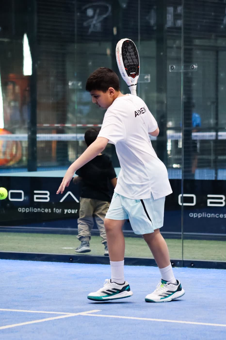 Junior padel player following through on a forehand, racket raised behind the shoulder, on a blue indoor court