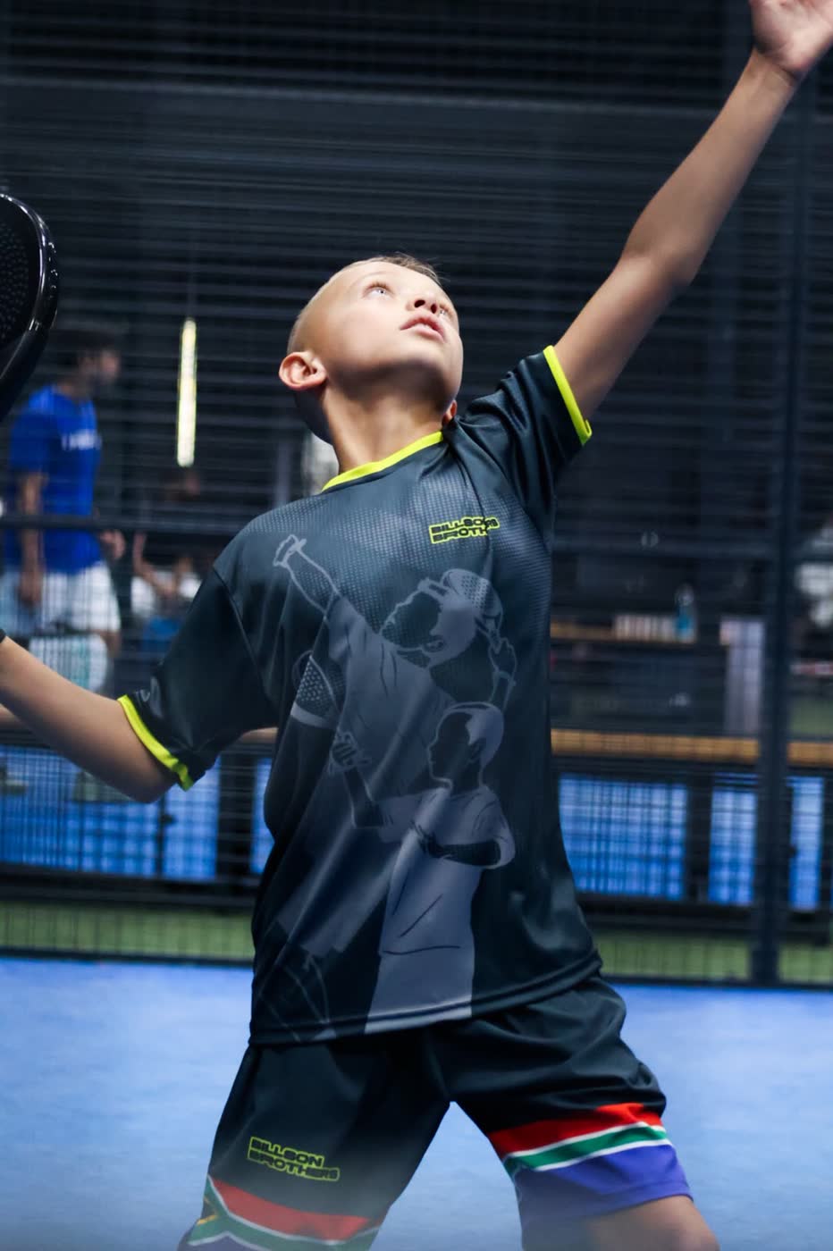 Junior padel player in South African colours looking up to track the ball, racket arm raised, on a blue indoor court