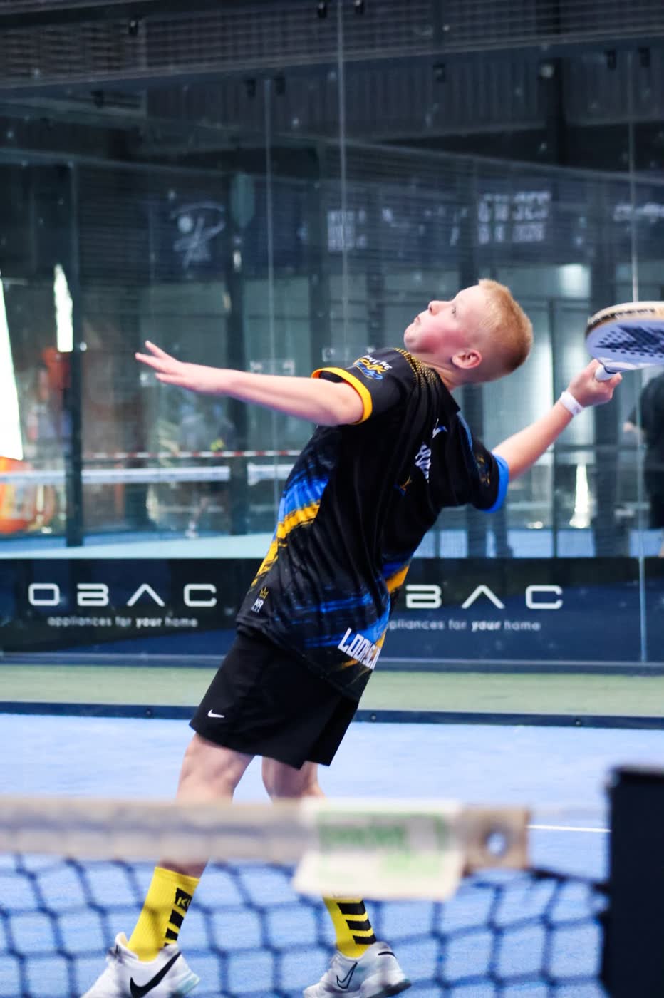 Junior padel player arching back to serve, eyes on the ball, in a blue and yellow team kit on an indoor court