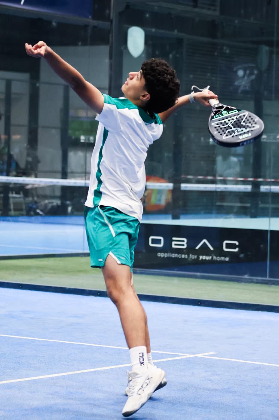 Junior padel player in teal and white kit stretching into an overhead serve, arm extended toward the ball