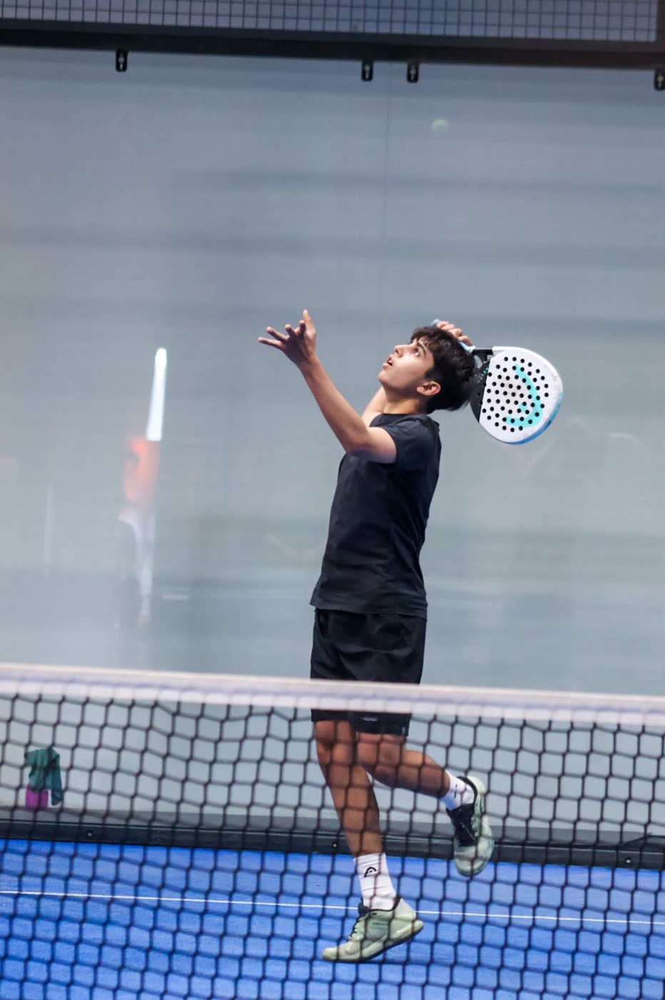 Junior padel player in black kit reaching overhead to strike a smash above the net
