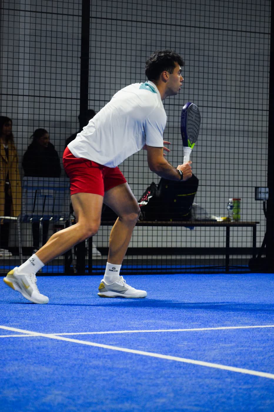 Padel player Leandro Augsburger crouched in a ready stance at the net, racket raised, on a blue indoor padel court