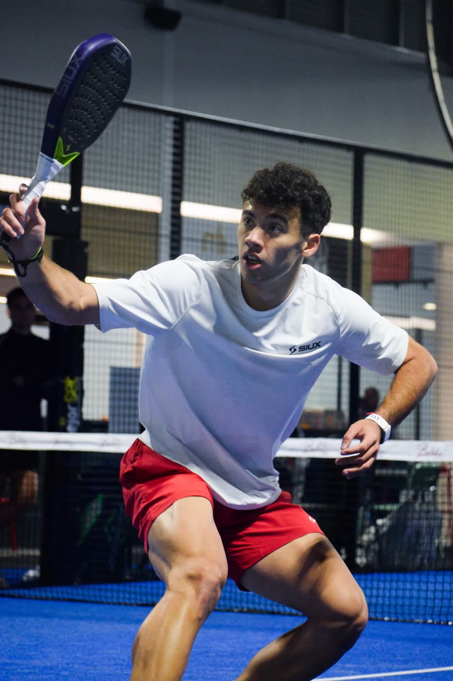 Padel player Leandro Augsburger mid-rally, racket drawn back for a forehand at the net on a blue indoor court